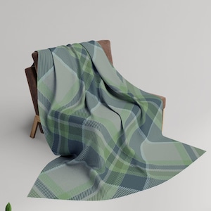 Può includere: Una morbida coperta con un motivo a quadri su larga scala nelle tonalità del verde salvia, verde bosco e blu ardesia. Il tessuto presenta una trama intrecciata con linee diagonali incrociate e blocchi di colore, drappeggiata su una sedia marrone.