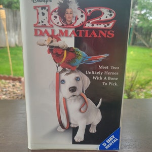 May include: A VHS tape cover for the movie 102 Dalmatians featuring Glenn Close as Cruella De Vil. The cover shows a dalmatian puppy with a red leash and a parrot with a yellow leash. The text on the cover reads "Disney's 102 Dalmatians" and "Meet Two Unlikely Heroes With A Bone To Pick."
