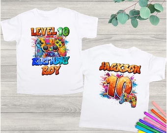 Könnte beinhalten: Zwei weiße T-Shirts mit farbenfrohen Grafiken zum Thema Videospiele. Ein Shirt trägt die Aufschrift "LEVEL 10 BIRTHDAY BOY" mit einer Controller-Grafik. Das andere Shirt trägt die Aufschrift "JACKSON 10" mit einer Controller-Grafik. Die Shirts befinden sich auf einem hellen Holzgrund.