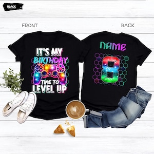 May include: Black t-shirts with birthday graphics. The front shirt says "IT'S MY BIRTHDAY TIME TO LEVEL UP" with a colorful video game controller. The back shirt has the word "NAME" and a number 8 in a hexagon pattern.