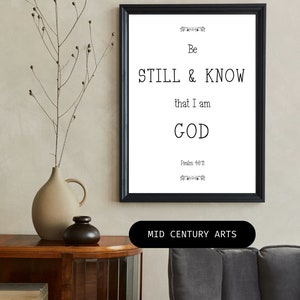 May include: Black and white framed print with the text "Be Still & Know that I am GOD Psalm 46:11" on a white background. The print is hanging on a beige wall above a wooden table with a vase and a small decorative object.