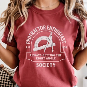 May include: A maroon t-shirt with a white graphic that reads "Protractor Enthusiast Society" with a protractor and compass illustration. The text "Always Getting The Right Angle" is below the illustration. The shirt is short-sleeved.