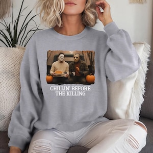 Könnte beinhalten: Hellgraues Sweatshirt mit einem Aufdruck von Michael Myers und Jason Voorhees, mit Kürbissen. Der Text "CHILLIN' BEFORE THE KILLING" ist darunter gedruckt. Ein humorvolles Halloween-Design.