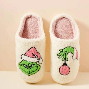 以下が含まれることがあります： 白くてふわふわのクリスマスをテーマにしたスリッパのペア。片方のスリッパには、ピンクのサンタ帽をかぶった緑色の顔、もう一方にはピンクのオーナメントを持った緑色の手があります。内側はソフトピンクです。