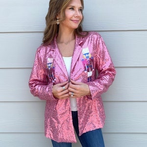 May include: A pink sequined blazer with a classic lapel collar and two nutcracker figures embroidered on the front. The jacket is open, revealing a white top underneath. The model is wearing blue jeans.