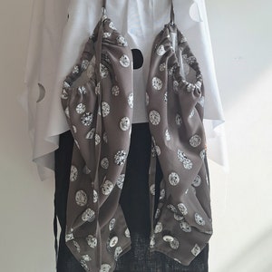 May include: A white poncho with circular cutouts layered over a black garment. Two grey fabric pouches with a white diamond pattern are suspended from the neck by thin straps, featuring gathered openings and white buttons at the bottom edges.