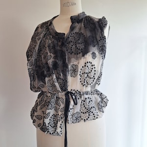Pode incluir: Uma blusa transparente sem mangas com um padrão floral preto e branco. A blusa apresenta uma cintura franzida com um laço preto e é exibida em um manequim. A parte superior tem um decote redondo e uma bainha com babados.