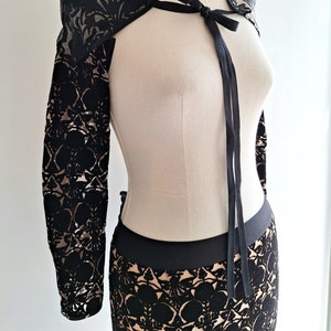 May include: Black lace bolero jacket with long sleeves and a matching skirt. The bolero features a delicate floral pattern and is tied at the front with a black ribbon. The skirt has a black waistband and a similar lace design.