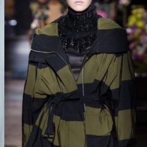 May include: A Dries Van Noten jacket featuring horizontal olive green and black stripes, a wide collar, a front zip closure, and a tie-waist detail. The garment is shown hanging on a hanger and worn on a runway, displaying a structured silhouette.
