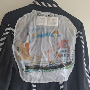 May include: A dark navy blue jacket featuring white and navy diagonal striped trim on the collar, sleeves, and hem. The back displays a large, circular white fabric patch with a printed illustration of a gondola in Venice and the text Moschino.