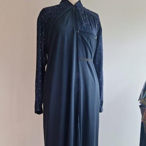 May include: A long, dark navy blue dress with long sleeves and a collared neckline. The sleeves have a dotted pattern, and the dress has a front opening. A black belt cinches the waist. The dress is displayed on a mannequin.