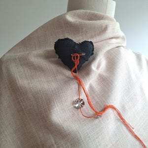 Puede incluir: Un broche negro en forma de corazón hecho de tela, con hilo naranja y una cuenta de cristal. El broche está sujeto a una tela de color claro, posiblemente lino, drapeada sobre un maniquí.