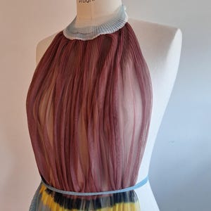 May include: A sheer, pleated halter dress in shades of burgundy, yellow, black, and blue. The dress features a light blue trim around the neck and waist. The dress is displayed on a mannequin.