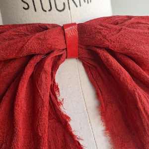 May include: A vibrant red scarf is held together by a matching leather band. The scarf has a textured appearance and frayed edges. The word "STOCKMAN" is printed on the mannequin.