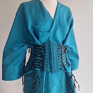 May include: A teal-colored wrap-style garment with wide sleeves and a V-neckline, featuring a structured black corset-style waist cincher with black ribbon lacing that crisscrosses down the front and ties at the bottom on both sides.