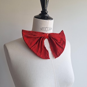 May include: A vibrant red bow tie made of textured fabric, displayed on a mannequin. The bow tie has a classic shape with a slightly distressed edge, adding a vintage feel. The mannequin is a neutral color, allowing the bow tie's color to stand out.