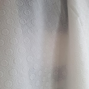 Pode incluir: Close-up de um tecido branco com um padrão floral e geométrico repetitivo. O tecido tem uma superfície texturizada com um brilho sutil, criando uma aparência suave e elegante. O design apresenta pequenos motivos florais detalhados dentro de formas hexagonais.