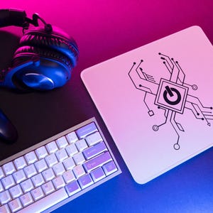Puede incluir: Un ratón blanco con un diseño de placa de circuito negro y un símbolo de botón de encendido. El ratón está sobre una superficie negra con un teclado blanco, un mando de juego negro y unos auriculares negros.