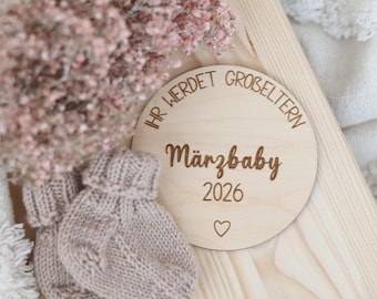 Pode incluir: Um disco de madeira com o texto em alemão "IHR WERDET GROBELTERN Märzbaby 2026" e um símbolo de coração. Ao lado, dois sapatinhos de bebé de malha em castanho claro. Flores cor-de-rosa no fundo.
