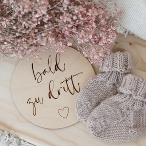 Könnte beinhalten: Ein Paar hellbraune gestrickte Babyschuhe neben einer Holzscheibe mit den Worten "bald zu dritt" und einem Herzsymbol. Rosa Blumen sind im Hintergrund. Die Schuhe sind aus weichem, strukturiertem Garn.