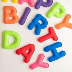 May include: A collection of individual, stuffed felt letters spelling out the phrase HAPPY BIRTHDAY. Each letter features a vibrant, solid color including yellow, pink, purple, lime green, orange, blue, and red, with visible stitched edges.