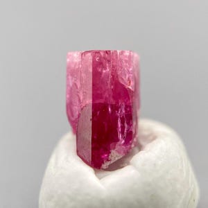 May include: A vibrant, translucent pink gemstone crystal with a distinct hexagonal prismatic structure. The mineral displays deep magenta hues, sharp vertical facets, and a natural, slightly irregular surface texture with visible internal inclusions.