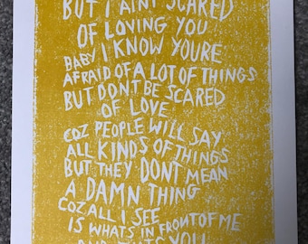May include: A yellow print with white text that reads "BABY I'M AFRAID OF A LOT OF THINGS BUT I AINT SCARED OF LOVING YOU..." The print is on white paper with a small white label in the corner with the number 1.