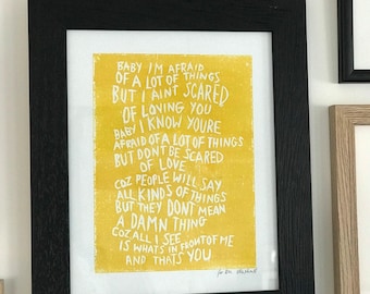 May include: A framed print with a yellow background and white handwritten text. The black frame surrounds the artwork, which features a quote about love. A small blue felt owl sits above the frame.