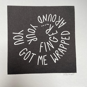 May include: A black square print with white text that reads "You Got Me Wrapped Around Your Finger". The text is arranged in a spiral pattern. The print is on a white background.
