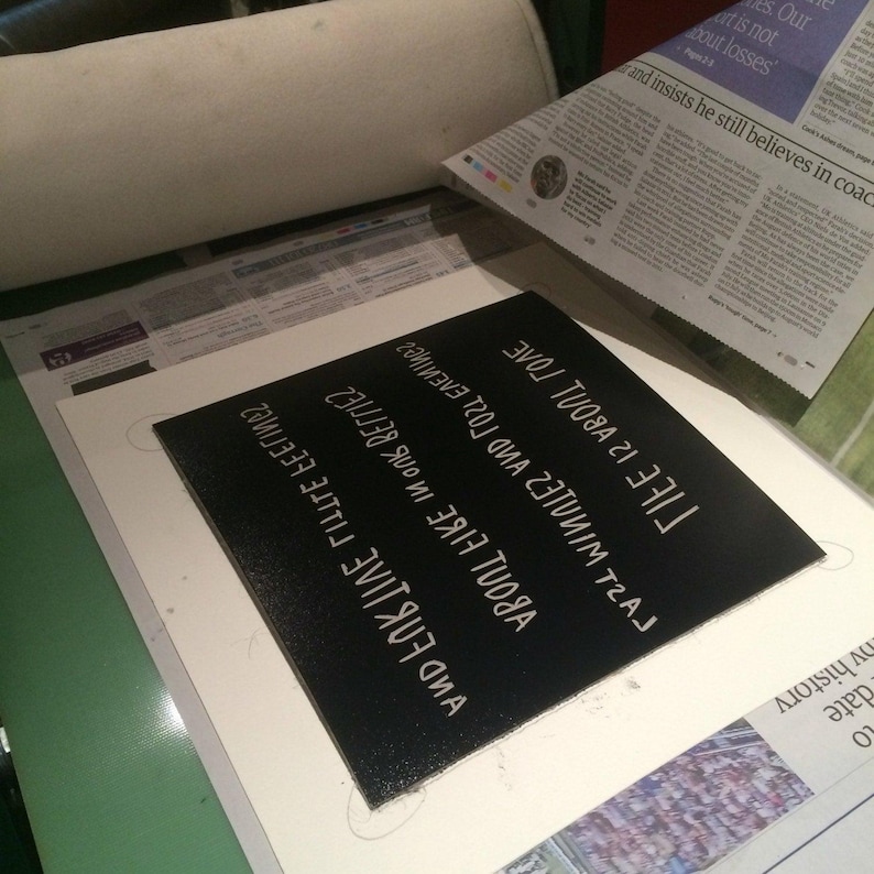 May include: A black square with white text that reads "And Fortune About Fire Little Better Last Minutes 231nim Traj Life Is About Love". The square is on a white surface, with newspaper and a roll of fabric in the background.