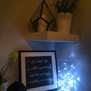 May include: A white shelf displays a geometric glass terrarium, a potted plant, and a framed print with the text "LIFE IS ABOUT LOVE." A glass vase with fairy lights and a small black lantern complete the scene.