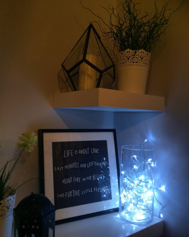 May include: A white shelf displays a geometric glass terrarium, a potted plant, and a framed print with the text "LIFE IS ABOUT LOVE." A glass vase with fairy lights and a small black lantern complete the scene.