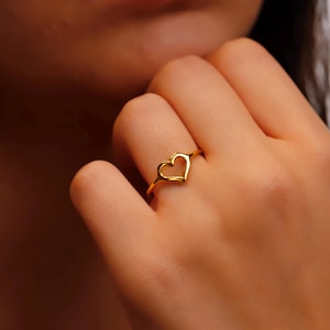 May include: A gold-colored ring featuring a heart-shaped design. The ring is worn on a finger, showcasing the delicate, open-heart silhouette. The ring's smooth, polished surface reflects light, highlighting its simple elegance.