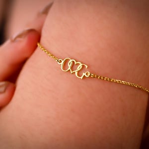 May include: A delicate gold bracelet with a chain link design. The bracelet features the intertwined letters "O" and "K" with a heart shape in the middle. The bracelet is worn on a wrist.