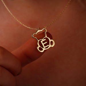 May include: Gold necklace with a teddy bear pendant featuring the letter E inside a heart.