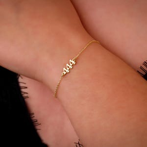 May include: A delicate gold-colored bracelet featuring the number "444" as a charm. The bracelet is on a slender chain and is worn on a wrist. The background is a blurred view of skin and black fabric.