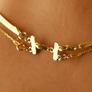 May include: Two gold necklaces layered on a person's neck. The top necklace is a thin, flat chain. The bottom necklace is a thicker chain with rectangular links.