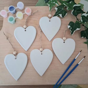 May include: Five white heart-shaped ornaments with twine hangers and wooden beads, displayed on a wooden board. Small paint pots in various colours and two blue-handled paintbrushes are also visible. Green and white ivy adds a decorative touch.