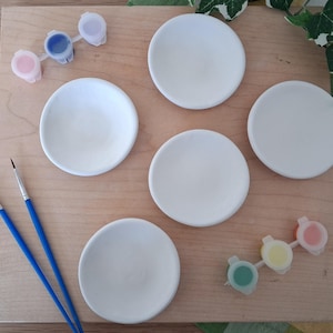 May include: A wooden board with five white ceramic coasters, small paint pots in pink, blue, and yellow, and two paintbrushes with blue handles. The coasters are ready to be painted.