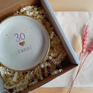 May include: A white ceramic dish with a gold rim, inscribed with the number "30", a heart, and the name "Sammi" in purple. The dish is nestled in a cardboard box with shredded paper, alongside dried flowers.