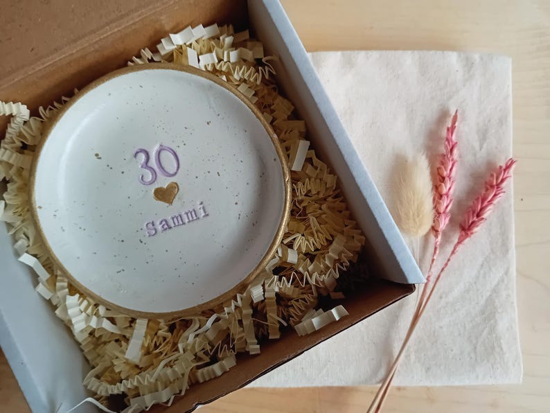 May include: A white ceramic dish with a gold rim, inscribed with the number "30", a heart, and the name "Sammi" in purple. The dish is nestled in a cardboard box with shredded paper, alongside dried flowers.
