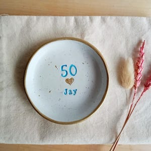 May include: A white ceramic plate with a gold rim, featuring the number "50", a heart, and the name "Jay" in blue. The plate rests on a beige linen cloth, with dried pink and beige flowers to the side. A decorative item for a birthday.