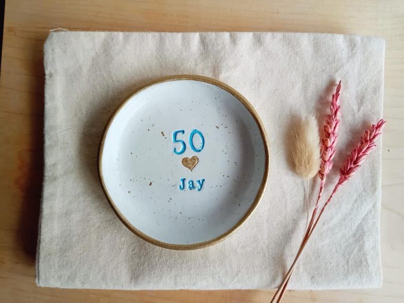 May include: A white ceramic plate with a gold rim, featuring the number "50", a heart, and the name "Jay" in blue. The plate rests on a beige linen cloth, with dried pink and beige flowers to the side. A decorative item for a birthday.