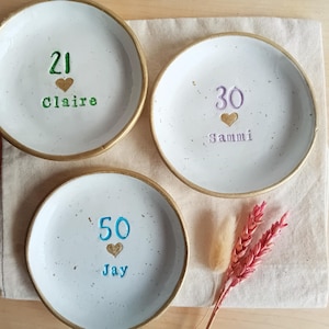 May include: Three white ceramic plates with gold rims, each inscribed with a number, a heart, and a name. The plates are arranged on a beige cloth, with decorative dried flowers. The numbers are 21, 30, and 50.