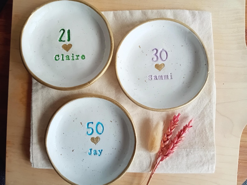 May include: Three white ceramic plates with gold rims, each inscribed with a number, a heart, and a name. The plates are arranged on a beige cloth, with decorative dried flowers. The numbers are 21, 30, and 50.