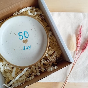 May include: A white ceramic dish with a gold rim, inscribed with the number "50", a heart, and the name "Jay" in blue. The dish is nestled in a cardboard box with paper filler, alongside a white cloth and pink dried flowers.