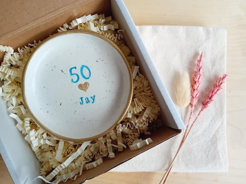 May include: A white ceramic dish with a gold rim, inscribed with the number "50", a heart, and the name "Jay" in blue. The dish is nestled in a cardboard box with paper filler, alongside a white cloth and pink dried flowers.