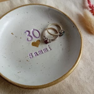 May include: A handmade ceramic ring dish with a gold rim, featuring the number "30", a gold heart, and the name "Sammi" in purple. Two rings rest inside the dish. The dish is white with speckled details.