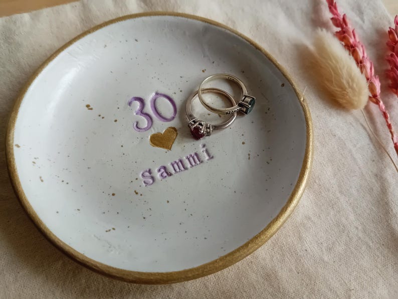 May include: A handmade ceramic ring dish with a gold rim, featuring the number "30", a gold heart, and the name "Sammi" in purple. Two rings rest inside the dish. The dish is white with speckled details.