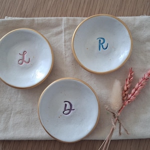 May include: Three small, white ceramic dishes with gold rims, each with a different initial in cursive: L (pink), R (blue), and D (purple). They sit on a beige linen cloth, with a small bunch of dried pink flowers.